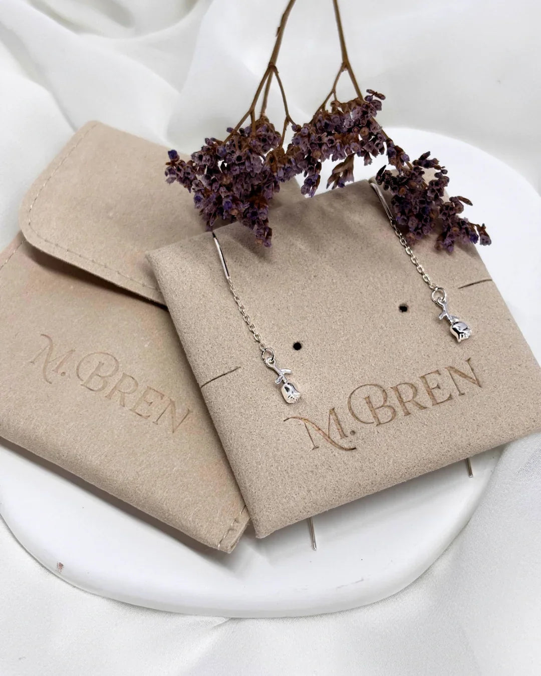 עגילי שרשרת עדינים מכסף סטרלינג עם תליון פרח דק, על רקע שקיות תכשיטים של M.Bren